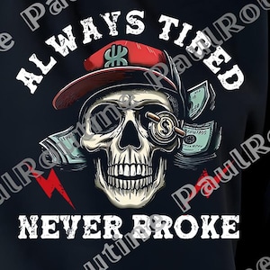 May include: Black sweatshirt with a graphic design featuring a skull wearing a red baseball cap and money. The text reads "ALWAYS TIRED NEVER BROKE" in white lettering. A red lightning bolt is also included in the design.