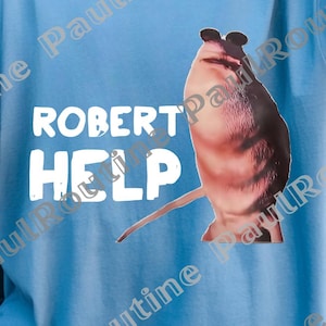 Könnte beinhalten: Hellblaues T-Shirt mit dem weißen Text "ROBERT HELP". Auf der Vorderseite ist eine Cartoon-Illustration einer Kreatur aufgedruckt. Das T-Shirt hat einen lässigen, übergroßen Stil.