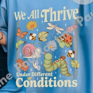 May include: Light blue t-shirt with the text "We All Thrive Under Different Conditions." The graphic features colorful illustrations of a snail, butterfly, ladybug, bee, caterpillar, and flowers. The shirt has a relaxed fit.