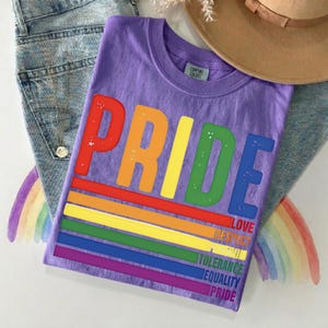 Peut inclure: Un t-shirt violet avec le mot "PRIDE" en grandes lettres aux couleurs de l'arc-en-ciel. En dessous, des rayures arc-en-ciel et les mots "LOVE, RESPECT, TOLERANCE, EQUALITY, PRIDE". Le t-shirt est sur un jean bleu et un chapeau marron.