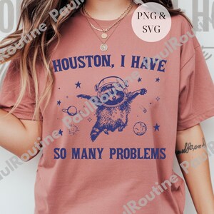 以下が含まれることがあります： 「Houston, I Have So Many Problems」のテキストが入ったダスティローズ色のTシャツ。宇宙ヘルメットをかぶったアライグマが星と惑星に囲まれているデザイン。カジュアルで着心地の良いスタイルです。