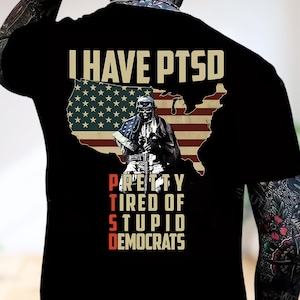Puede incluir: Camiseta negra con un diseño gráfico que presenta el texto "I HAVE PTSD" sobre un contorno de la bandera estadounidense y una ilustración de un soldado. Debajo de la bandera, el texto dice "PRETTY TIRED OF STUPID DEMOCRATS."