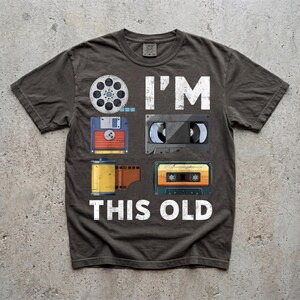 Könnte beinhalten: Dunkelgraues T-Shirt mit dem Text "I'M THIS OLD" und Grafiken einer Filmrolle, Diskette, Filmrolle, VHS-Kassette und Audiokassette. Das Shirt hat ein Retro-Design.