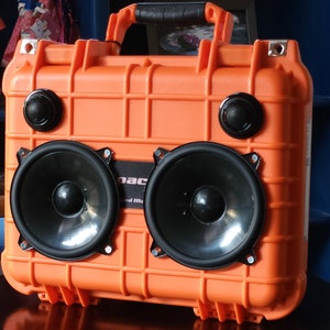 Puede incluir: Caja naranja impermeable con dos altavoces grandes y dos más pequeños. La caja tiene un asa negra y el texto "pac" y "and music" es visible en la parte delantera.