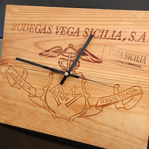Puede incluir: Reloj de pared de madera con manecillas negras, con el texto "BODEGAS VEGA SICILIA, S.A." y un logotipo. La esfera del reloj es de madera clara con detalles grabados, incluyendo las palabras "VEGA SICILIA" y "TISSERVA ESPECIAL".