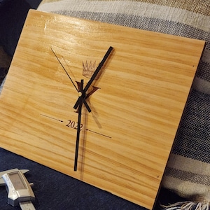 May include: Square wooden clock with a light brown finish. The clock face features black hands, a crown graphic, and the year "2022" printed below the centre. A silver caliper is in the foreground.