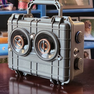 Può includere: Un altoparlante portatile nero, robusto, con due grandi altoparlanti e una maniglia. L'altoparlante ha più porte e pulsanti sul lato.