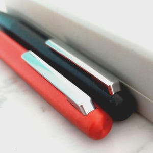 May include: Close-up of two pens, one red and one black, with silver clips. The pens are positioned diagonally, with a white rectangular object in the background. The red pen is in the foreground.