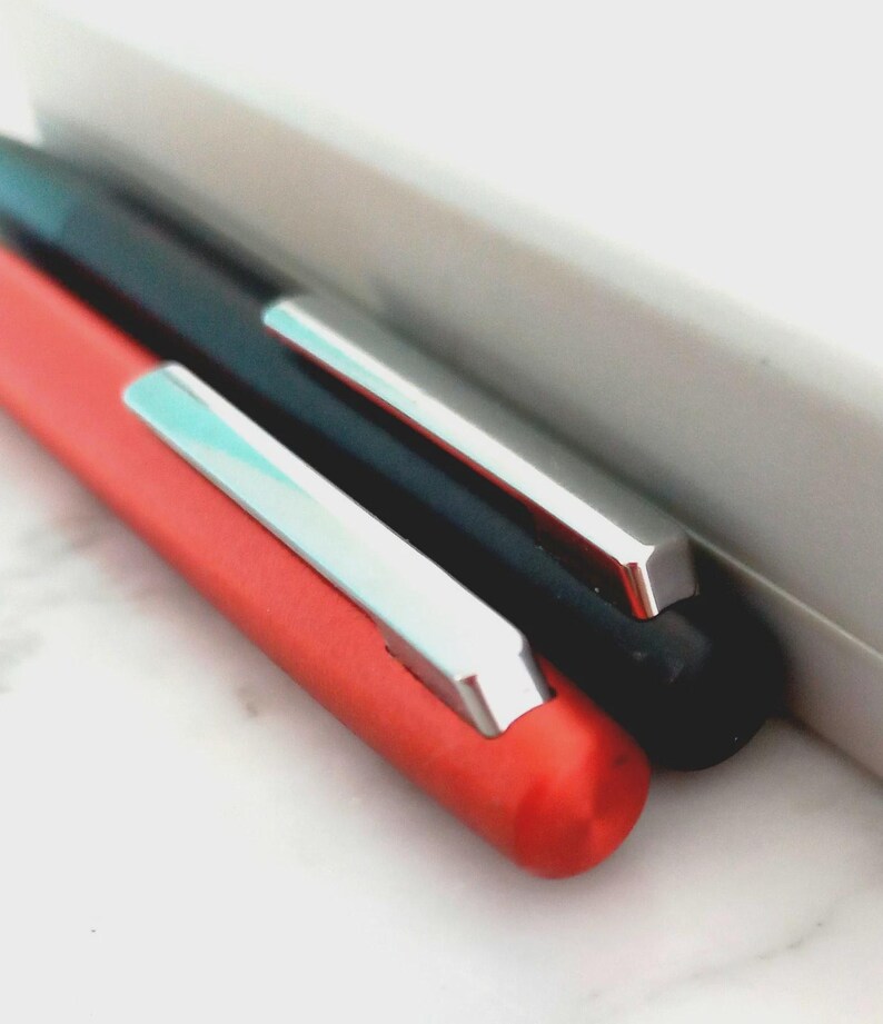 May include: Close-up of two pens, one red and one black, with silver clips. The pens are positioned diagonally, with a white rectangular object in the background. The red pen is in the foreground.