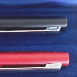 May include: Close-up of two Lamy pens. One pen is black, the other is red. Both pens have a silver clip with the Lamy brand name. The pens are set against a blue background.