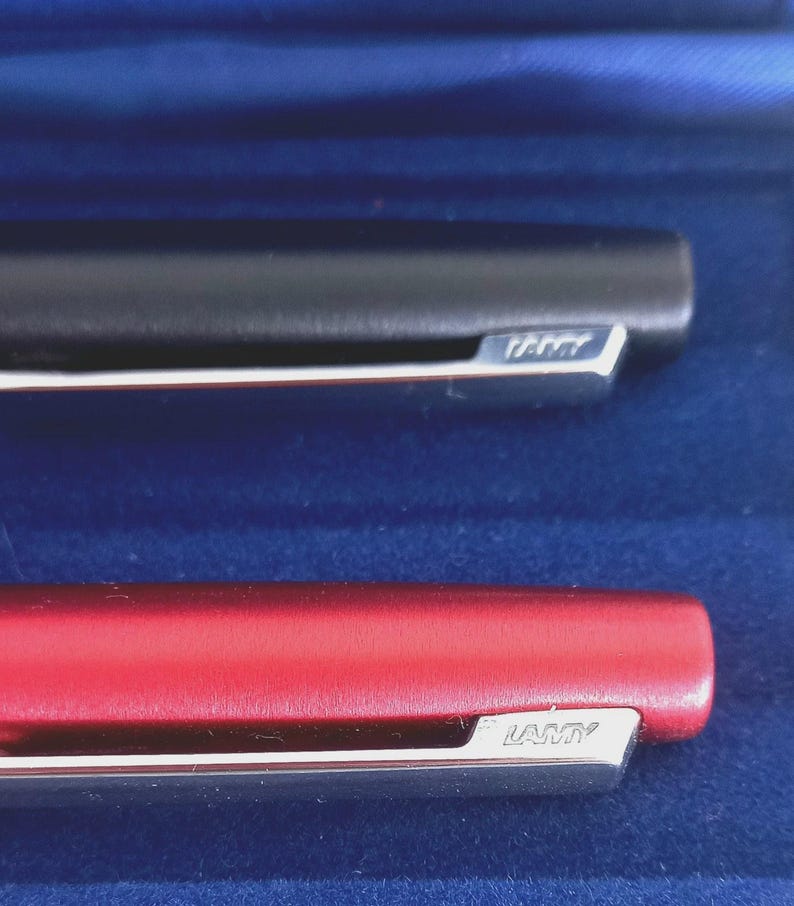 May include: Close-up of two Lamy pens. One pen is black, the other is red. Both pens have a silver clip with the Lamy brand name. The pens are set against a blue background.