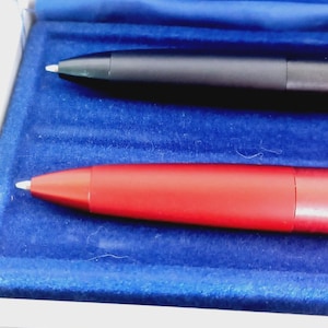 May include: Two pens, one black and one red, are displayed in a blue velvet-lined box. The pens have a sleek, modern design with silver tips. The red pen is a vibrant, solid color, while the black pen has a matte finish.