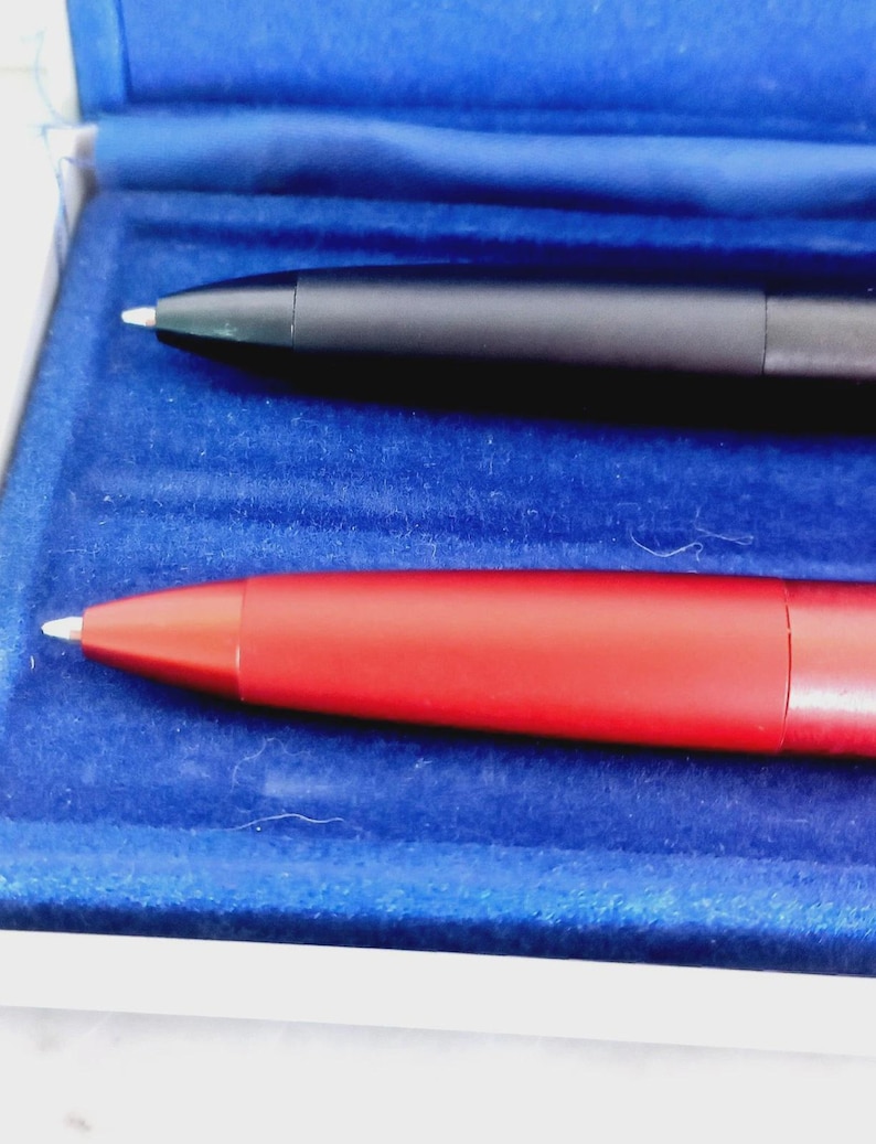 May include: Two pens, one black and one red, are displayed in a blue velvet-lined box. The pens have a sleek, modern design with silver tips. The red pen is a vibrant, solid color, while the black pen has a matte finish.