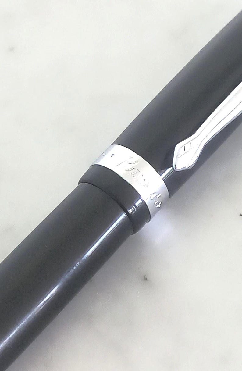 K&ouml;nnte beinhalten: Nahaufnahme eines schwarzen F&uuml;llfederhalters mit silbernem Band. Der Stift hat eine glatte, gl&auml;nzende Oberfl&auml;che und einen silbernen Clip. Das Band hat gravierten Text. Der Stift wird wahrscheinlich zum Schreiben und f&uuml;r Kalligraphie verwendet.
