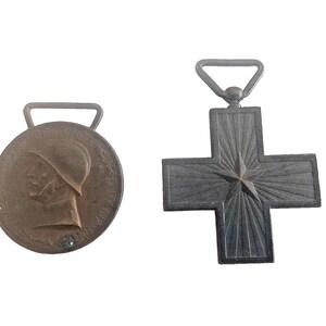 May include: Two antique medals. One is a bronze circular medal with a profile of a person wearing a helmet. The other is a dark grey cross-shaped medal with a star in the centre.