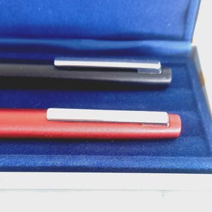 May include: Close-up of two pens in a blue velvet box. One pen is black, the other is red. Both pens have a silver clip. The pens are displayed in a white box with a blue velvet interior.