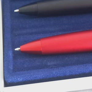 May include: Close-up of two pens in a presentation box. One pen is black, the other is red. Both pens have a white tip. The pens are set in a blue velvet-lined box with a white border.