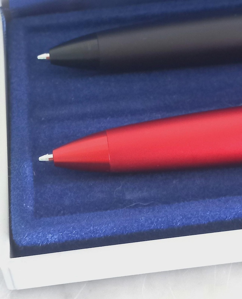 May include: Close-up of two pens in a presentation box. One pen is black, the other is red. Both pens have a white tip. The pens are set in a blue velvet-lined box with a white border.