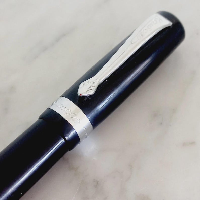 K&ouml;nnte beinhalten: Nahaufnahme eines schwarzen F&uuml;llfederhalters mit silbernem Clip und Akzenten. Der Clip ist mit dem Markennamen und einem stilisierten Design graviert. Der Stift hat eine gl&auml;nzende Oberfl&auml;che.