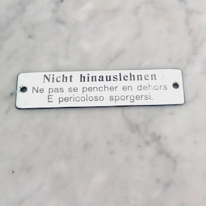 Puede incluir: Un letrero rectangular de metal blanco con esquinas redondeadas y dos agujeros. El letrero tiene texto negro en tres líneas: "Nicht hinauslehnen / Ne pas se pencher en dehors / E pericoloso sporgersi."