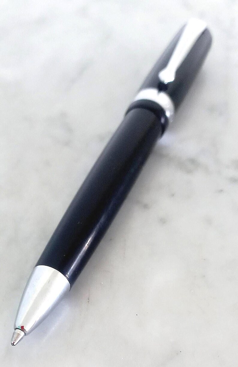 K&ouml;nnte beinhalten: Ein schwarzer Kugelschreiber mit silbernen Akzenten. Der Stift hat eine silberne Spitze und einen silbernen Clip. Er liegt auf einer wei&szlig;en Marmoroberfl&auml;che. Der Stift ist ein Schreibger&auml;t.