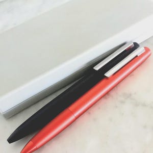 May include: Two pens, one black and one red, with silver clips, are positioned diagonally on a marble surface. A white rectangular box is in the background. The pens have a sleek, modern design.