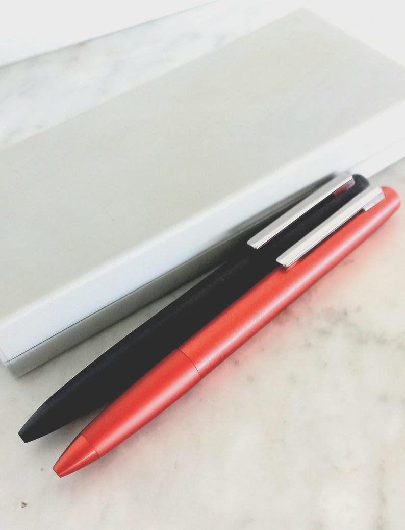 May include: Two pens, one black and one red, with silver clips, are positioned diagonally on a marble surface. A white rectangular box is in the background. The pens have a sleek, modern design.