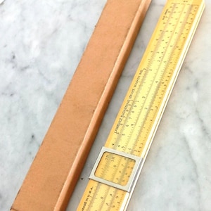 Może przedstawiać: Zabytkowa suwmiarka z beżowym korpusem i srebrną metalową ramą, obok brązowego kartonowego etui. Suwak ma oznaczenia pomiarowe oraz tekst "MADE IN U.S.A." i "LAWRENCE ENGINEERING SERVICE WABASH IND."