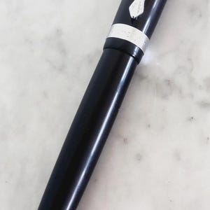 K&ouml;nnte beinhalten: Ein schwarzer Kugelschreiber mit silbernen Akzenten. Der Stift hat einen gl&auml;nzenden schwarzen Schaft, eine silberne Spitze und einen silbernen Clip. Er liegt auf einer wei&szlig;en Marmoroberfl&auml;che.