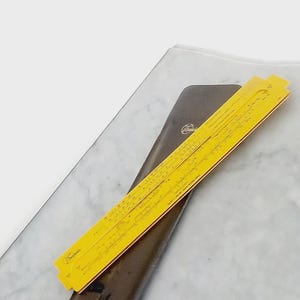 Puede incluir: Una regla de cálculo amarilla vintage con bordes rojos descansa sobre un estuche de cuero marrón oscuro. La regla de cálculo es una herramienta clásica para cálculos matemáticos. El estuche está grabado con un logotipo. Los artículos están sobre una superficie de mármol.