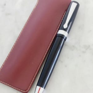 K&ouml;nnte beinhalten: Ein schwarzer Stift mit silbernen Akzenten und einer rot-silbernen Spitze, neben einem weinroten Leder-Stiftetui. Der Stift hat einen silbernen Clip und ein silbernes Band. Das rechteckige Etui ist wei&szlig; gen&auml;ht.