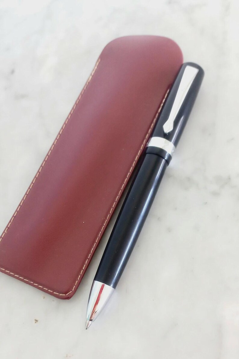 K&ouml;nnte beinhalten: Ein schwarzer Stift mit silbernen Akzenten und einer rot-silbernen Spitze, neben einem weinroten Leder-Stiftetui. Der Stift hat einen silbernen Clip und ein silbernes Band. Das rechteckige Etui ist wei&szlig; gen&auml;ht.