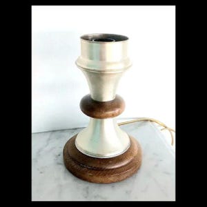 Può includere: Una base per lampada vintage con una parte superiore in metallo color argento e una base in legno. La lampada ha un design classico con un portalampada nella parte superiore e un cavo di alimentazione. La base è in legno marrone scuro e ha una forma rotonda.