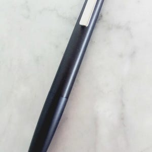 May include: A sleek, dark blue pen with a silver clip. The pen has a streamlined design and a pointed tip. The pen is lying on a white marble surface. The pen is approximately 5.5 inches long.