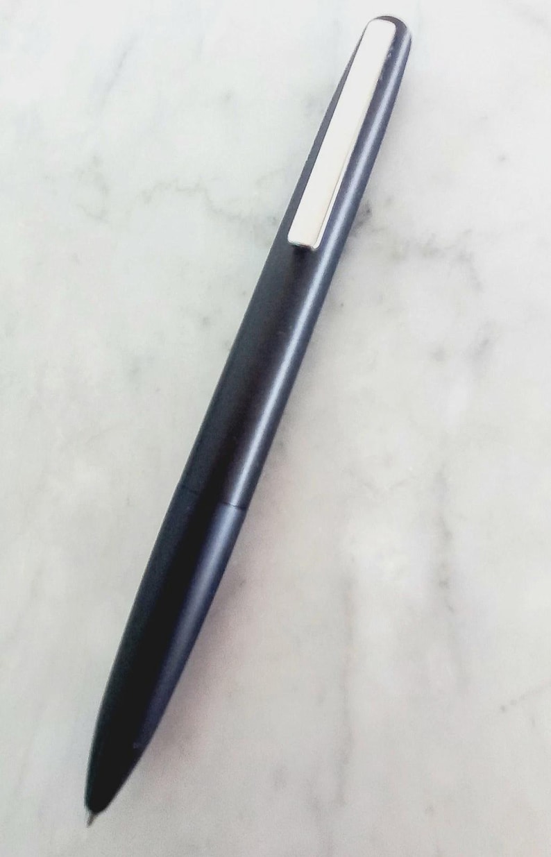 May include: A sleek, dark blue pen with a silver clip. The pen has a streamlined design and a pointed tip. The pen is lying on a white marble surface. The pen is approximately 5.5 inches long.