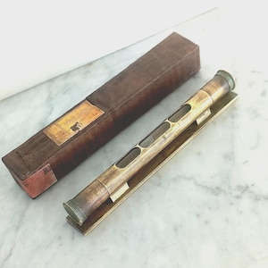 May include: An antique brass spirit level with a rectangular shape and multiple viewing windows. The level is resting on a brass base, next to a brown rectangular case with a label that reads "Spirit Level".