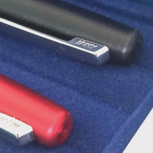 May include: Close-up of two pens, one black and one red, resting on a blue surface. The black pen has a silver clip with the brand name. The red pen is partially visible, also with a silver clip. The pens appear to be high-quality writing instruments.