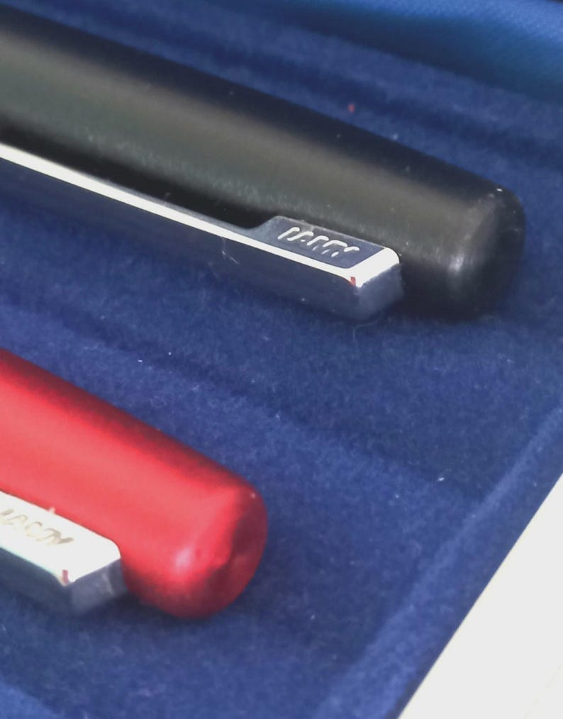 May include: Close-up of two pens, one black and one red, resting on a blue surface. The black pen has a silver clip with the brand name. The red pen is partially visible, also with a silver clip. The pens appear to be high-quality writing instruments.