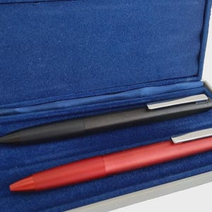 May include: A black and a red pen with silver clips rest inside a blue velvet-lined box. The pens are sleek and modern, suggesting a professional or luxury writing instrument.