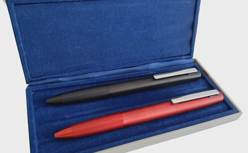 May include: A black and a red pen with silver clips rest inside a blue velvet-lined box. The pens are sleek and modern, suggesting a professional or luxury writing instrument.
