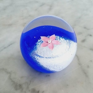 Puede incluir: Un pisapapeles de cristal transparente con un diseño azul y blanco. Una flor rosa está incrustada en el centro del pisapapeles. El pisapapeles es redondo y se encuentra sobre una superficie de mármol.