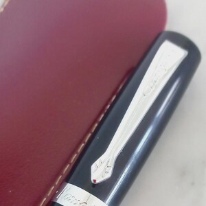 K&ouml;nnte beinhalten: Ein schwarzer F&uuml;llfederhalter mit silbernem Clip und Akzenten, neben einem weinroten Lederetui. Der Stift tr&auml;gt den Markennamen "Kaweco" eingraviert. Ein elegantes Schreibger&auml;t.