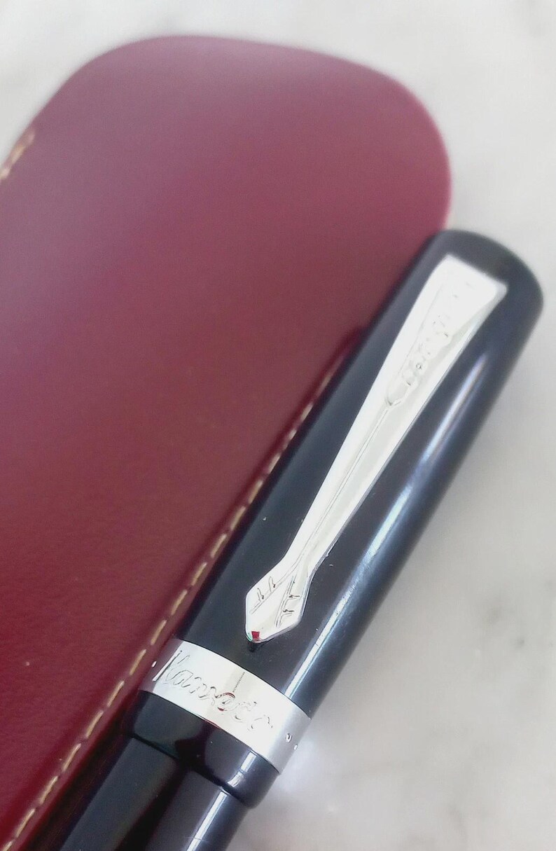 K&ouml;nnte beinhalten: Ein schwarzer F&uuml;llfederhalter mit silbernem Clip und Akzenten, neben einem weinroten Lederetui. Der Stift tr&auml;gt den Markennamen "Kaweco" eingraviert. Ein elegantes Schreibger&auml;t.