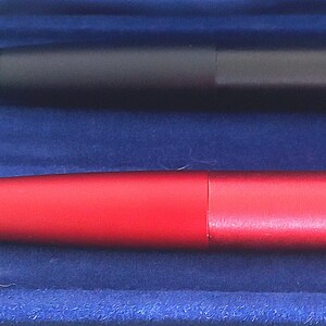 May include: Close-up of two pens. One pen is a matte black, and the other is a vibrant red. Both pens have a sleek, cylindrical design. The pens are set against a deep blue background, creating a striking contrast.