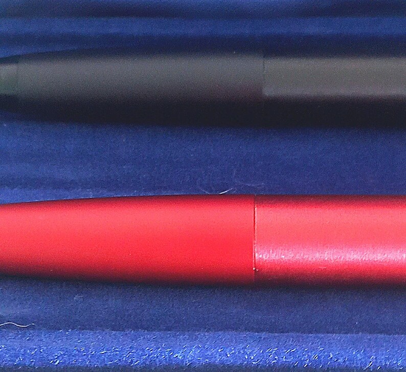 May include: Close-up of two pens. One pen is a matte black, and the other is a vibrant red. Both pens have a sleek, cylindrical design. The pens are set against a deep blue background, creating a striking contrast.