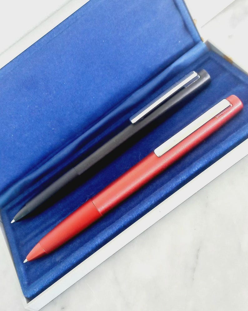 May include: Two pens, one black and one red, are displayed inside a blue velvet-lined box. Each pen has a silver clip. The red pen is in the foreground, and the black pen is behind it.