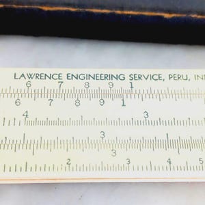 May include: Close-up of a vintage slide rule with the text "LAWRENCE ENGINEERING SERVICE, PERU, IND" printed on it. The rule has a cream-colored background with black markings and numbers. The numbers are clearly visible, indicating its use for calculations.
