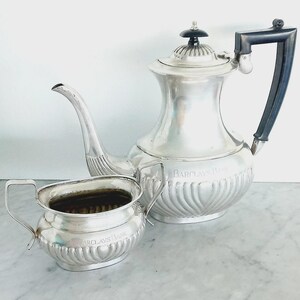 May include: A silver tea set, including a teapot and a creamer. The teapot has a black handle and a decorative lid. The creamer has two handles. Both pieces are engraved with the words "Barclays Bank".
