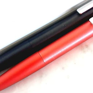 May include: Close-up of two pens. One pen is black, and the other is red. Both pens have a sleek, modern design with a metallic finish. The pens are lying diagonally across a white surface.