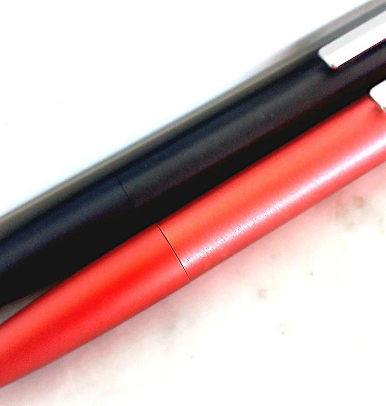 May include: Close-up of two pens. One pen is black, and the other is red. Both pens have a sleek, modern design with a metallic finish. The pens are lying diagonally across a white surface.